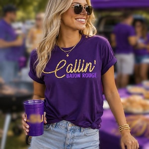 Puede incluir: Una camiseta morada de manga corta con el texto Callin' Baton Rouge en una fuente dorada con purpurina. La camiseta tiene cuello redondo y se combina con pantalones cortos de mezclilla azul claro. Se sostiene un vaso de pl&aacute;stico morado con logo dorado.