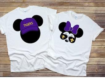 Könnte beinhalten: Zwei weiße T-Shirts mit einem Mickey Mouse und Minnie Mouse Design. Das Mickey Mouse Shirt hat einen lila Hut mit dem Wort "TIGERS" in gelben Buchstaben. Das Minnie Mouse Shirt hat eine lila Schleife und eine Sonnenbrille mit goldenen Fleur-de-lis.