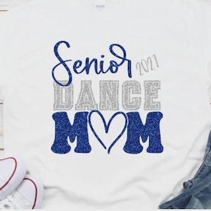 May include: A white short-sleeved t-shirt featuring the text Senior 2021 DANCE MOM in a glittery finish. The word Senior is in blue script, DANCE is in silver block letters, and MOM is in blue block letters with a heart replacing the letter O.