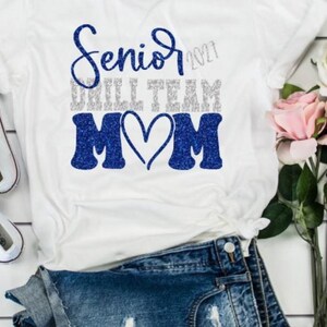 May include: A white t-shirt featuring blue and silver glitter text that reads Senior 2021 Drill Team Mom with a heart shape between the letters M and M. The shirt is displayed with white canvas sneakers, blue denim shorts, and pink roses.