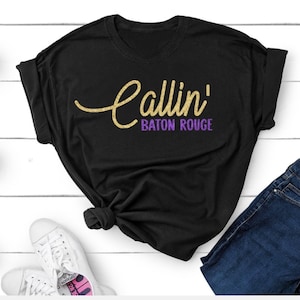 May include: A black short-sleeved t-shirt featuring the text Callin' in a gold glitter script font and Baton Rouge in a purple block font. The shirt is styled with a knot at the bottom hem and is paired with dark blue denim jeans and white sneakers.