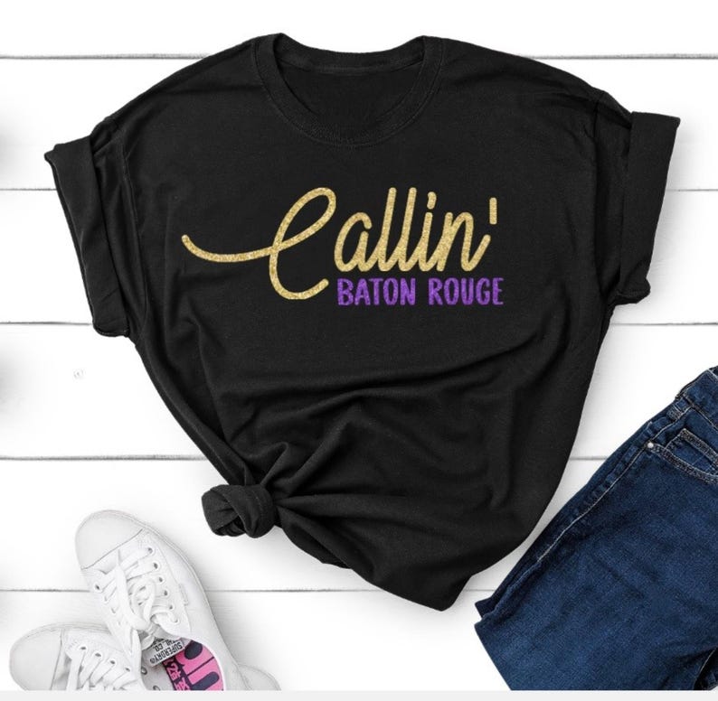 May include: A black short-sleeved t-shirt featuring the text Callin' in a gold glitter script font and Baton Rouge in a purple block font. The shirt is styled with a knot at the bottom hem and is paired with dark blue denim jeans and white sneakers.