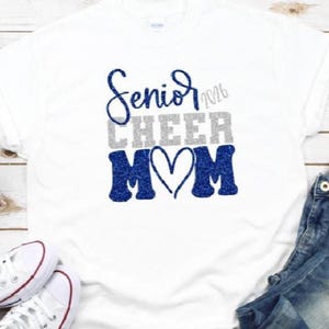 May include: White t-shirt with the text "Senior 2026 CHEER MOM" in blue and silver glitter. The shirt is paired with blue jeans and white sneakers with red accents, all on a light wood background.