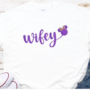 May include: White t-shirt with the word "wifey" in purple glitter script, accompanied by a purple Minnie Mouse bow design. The shirt is displayed with blue jeans and white sneakers.