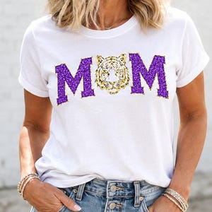 May include: A white short-sleeved t-shirt featuring the text MOM in purple glitter letters, with a tiger face graphic replacing the letter O. The shirt has a crew neckline and is paired with light blue denim shorts with a button-front closure.