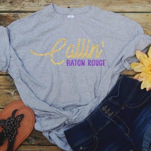 May include: A black short-sleeved t-shirt featuring the text Callin' in a gold glitter script font and Baton Rouge in a purple block font. The shirt is styled with a knot at the bottom hem and is paired with dark blue denim jeans and white sneakers.
