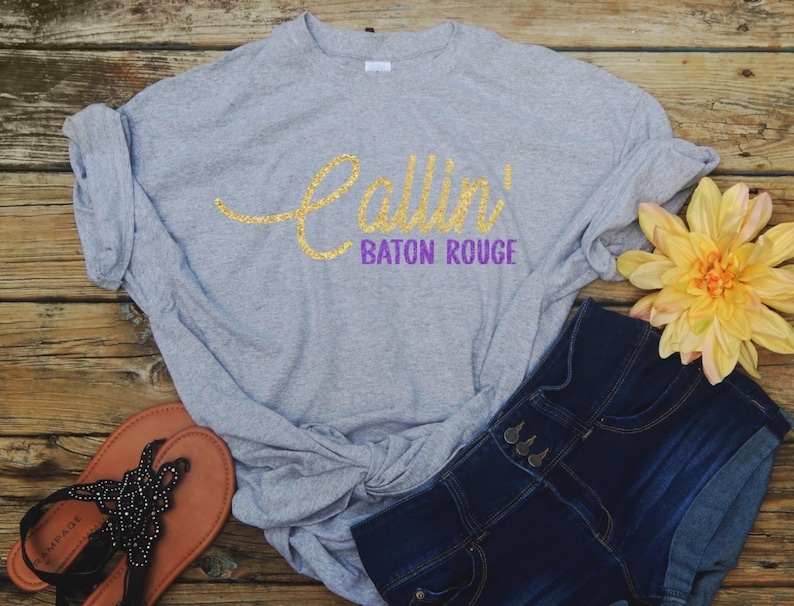 May include: A black short-sleeved t-shirt featuring the text Callin' in a gold glitter script font and Baton Rouge in a purple block font. The shirt is styled with a knot at the bottom hem and is paired with dark blue denim jeans and white sneakers.