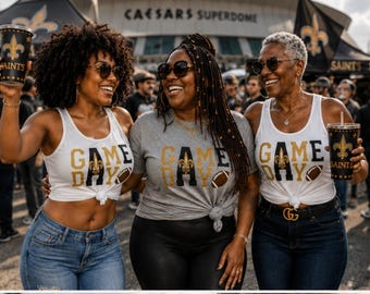 Puede incluir: Tres personas visten camisetas con el texto GAME DAY y un gráfico de flor de lis. Dos llevan camisetas de tirantes blancas con vaqueros azules, y una lleva una camiseta gris con pantalones negros. Sostienen vasos negros con logotipos dorados.