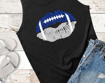 Puede incluir: Una camiseta sin mangas negra con un gráfico de labios diseñado para parecer un balón de fútbol americano. El labio superior es de purpurina azul con cordones blancos y el labio inferior es de purpurina plateada, centrado en la parte frontal.