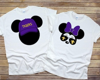 Puede incluir: Dos camisetas blancas con un diseño de Mickey Mouse morado y dorado. Una camiseta presenta a Mickey Mouse con un sombrero morado con la palabra "TIGERS" en letras amarillas. La otra camiseta presenta a Minnie Mouse con gafas de sol moradas con detalles dorados en forma de flor de lis.