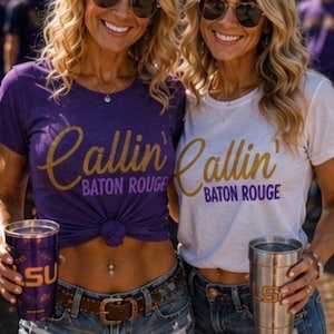 Puede incluir: Dos camisetas con el texto Callin' Baton Rouge en dorado y morado. Una camiseta es morada con texto dorado y la otra es blanca con texto dorado y morado. Ambas se combinan con pantalones cortos de mezclilla desgastados y vasos de metal.
