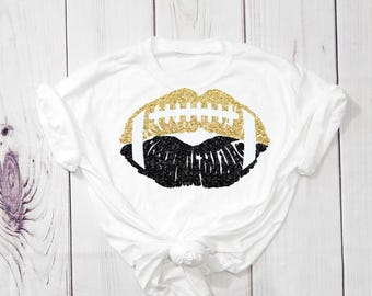 Puede incluir: Una camiseta blanca de manga corta con un gráfico central de labios con forma de balón de fútbol americano. La mitad superior de los labios es de purpurina dorada, la mitad inferior de purpurina negra, con cordones blancos en el centro.