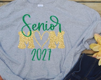 May include: A light grey t-shirt featuring the green glitter text Senior, the gold glitter text MOM with a heart shape in the centre, and the green glitter text 2027. The shirt has short sleeves and a crew neckline with a casual, relaxed fit.