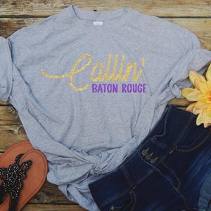 Puede incluir: Una camiseta gris jaspeado de manga corta con el texto Callin' en una fuente cursiva de purpurina dorada y BATON ROUGE en letras may&uacute;sculas moradas. La camiseta se muestra junto a unos pantalones cortos de mezclilla azul oscuro, sandalias marrones y una flor amarilla.