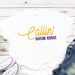 Puede incluir: Una camiseta blanca de manga corta con el texto Callin' en una fuente cursiva amarilla y el texto BATON ROUGE en una fuente sans-serif morada centrada en el pecho. La camiseta tiene un cuello redondo y un dise&ntilde;o de ajuste est&aacute;ndar.