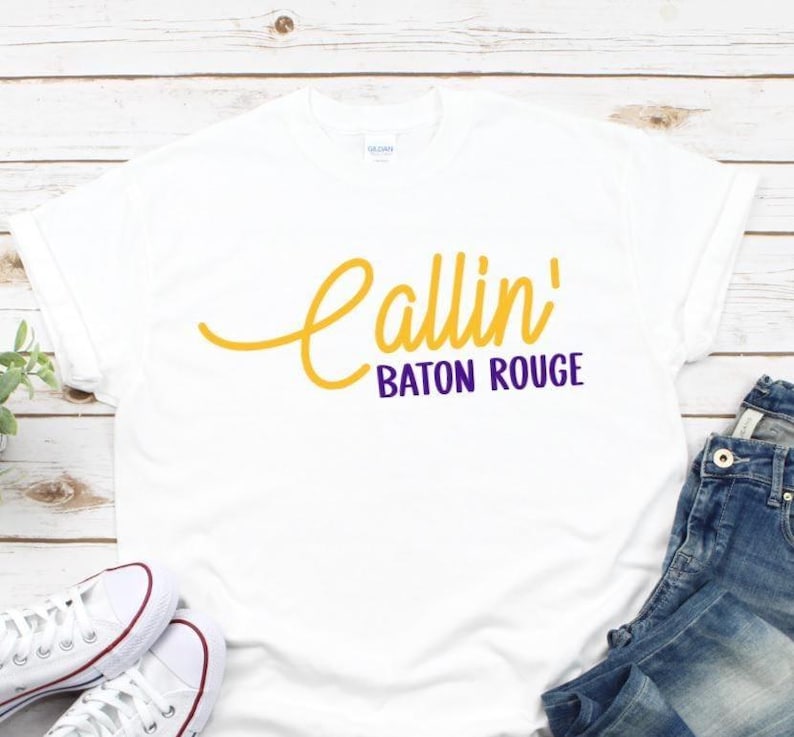 Puede incluir: Una camiseta blanca de manga corta con el texto Callin' en una fuente cursiva amarilla y el texto BATON ROUGE en una fuente sans-serif morada centrada en el pecho. La camiseta tiene un cuello redondo y un dise&ntilde;o de ajuste est&aacute;ndar.