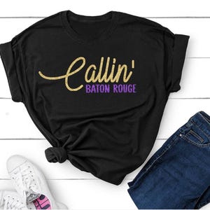 May include: A white short-sleeved t-shirt featuring the text Callin' in a yellow cursive script font and the words BATON ROUGE in a purple sans-serif font printed across the center of the chest area. The shirt has a standard crew neckline.