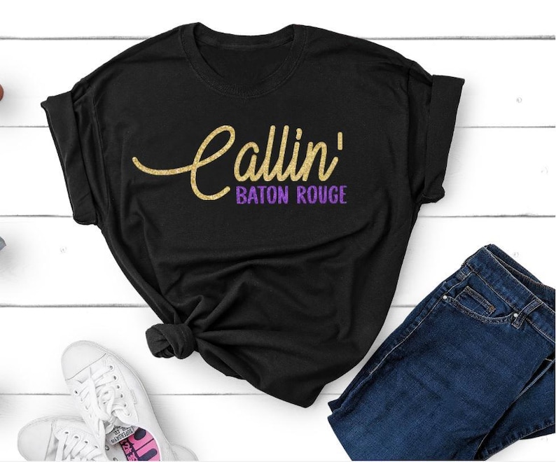 May include: A white short-sleeved t-shirt featuring the text Callin' in a yellow cursive script font and the words BATON ROUGE in a purple sans-serif font printed across the center of the chest area. The shirt has a standard crew neckline.