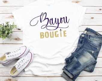 May include: White t-shirt with the words "Bayou Bougie" in purple script and gold glitter lettering. The shirt is paired with blue jeans and white sneakers. A small green plant is also in the image.