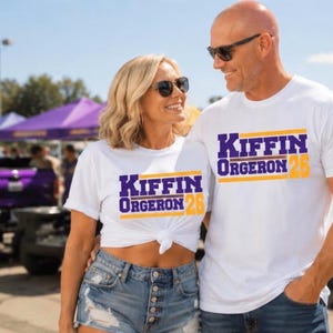 May include: Two white short-sleeved t-shirts featuring the purple and gold text "KIFFIN ORGERON 26" printed on the chest. The shirts are worn with blue denim shorts and blue jeans, respectively, and both individuals are wearing dark sunglasses.