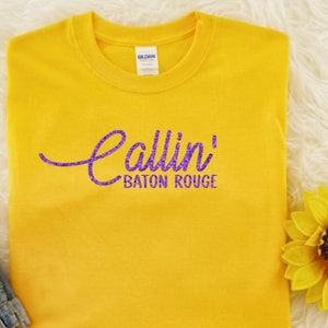 May include: A YELLOW short-sleeved t-shirt featuring the text Callin' in a purple cursive script font and the words BATON ROUGE in a purple sans-serif font printed across the center of the chest area. The shirt has a standard crew neckline.
