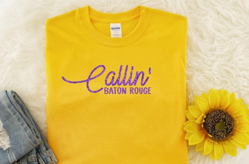 May include: A YELLOW short-sleeved t-shirt featuring the text Callin' in a purple cursive script font and the words BATON ROUGE in a purple sans-serif font printed across the center of the chest area. The shirt has a standard crew neckline.