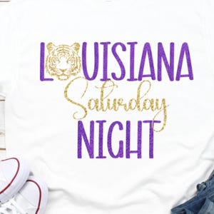 May include: A white short-sleeved t-shirt featuring a graphic print with the text LOUISIANA in purple, a gold glitter tiger head icon, the word Saturday in gold script, and the word NIGHT in purple, all displayed in a bold, glittery font style.