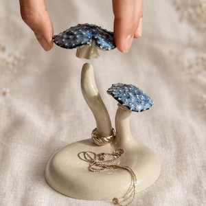 Puede incluir: Un joyero de cerámica con forma de hongo con dos hongos con gorro azul y una base color crema. Una mano sostiene uno de los sombreros de hongo. Un collar y un anillo plateados están en el soporte.