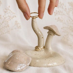 Puede incluir: Un soporte para anillos de cerámica con forma de setas. El soporte es de color blanco roto con motas marrones y presenta dos sombreros de setas y una pequeña base redonda. Un anillo dorado está colocado en el soporte.