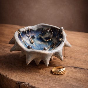 Op de afbeelding: Een keramische sieradenschaal met een blauw-grijze glazuur, met gouden kettingen en ringen. De schaal heeft een stekelige, stervormige vorm. Een gouden ring ligt op het houten oppervlak naast de schaal. De sieraden worden op een houten tafel getoond.