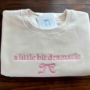 May include: A cream-colored sweatshirt with the text "a little bit dramatic" embroidered in pink. A pink bow is embroidered below the text.