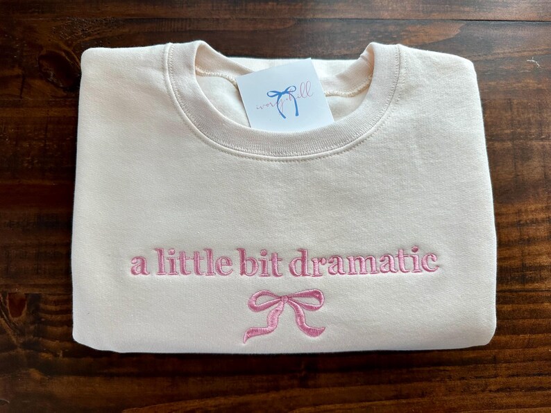 May include: A cream-colored sweatshirt with the text "a little bit dramatic" embroidered in pink. A pink bow is embroidered below the text.