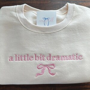 May include: A cream-colored sweatshirt with the text "a little bit dramatic" embroidered in pink. A pink bow is embroidered below the text.