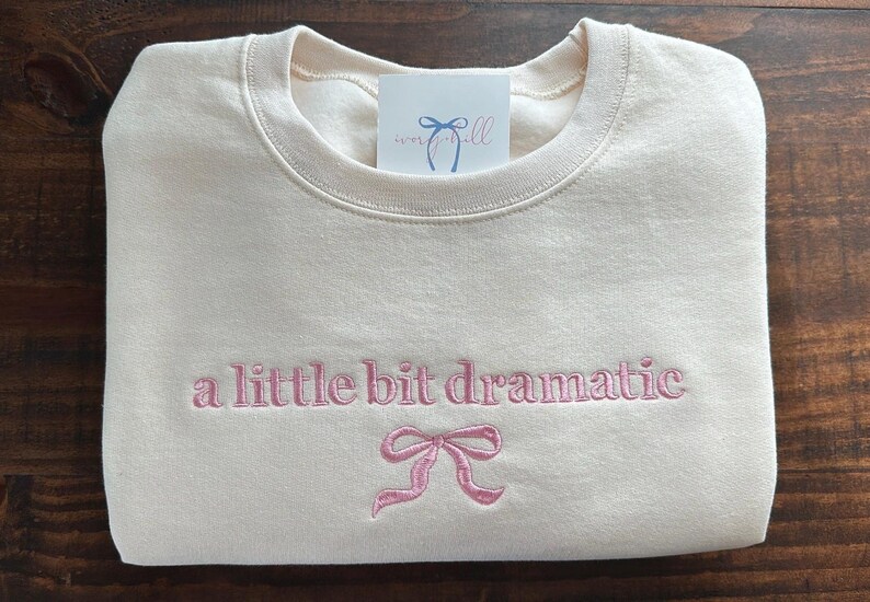 May include: A cream-colored sweatshirt with the text "a little bit dramatic" embroidered in pink. A pink bow is embroidered below the text.