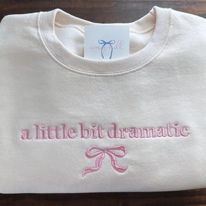May include: A cream-colored sweatshirt with the text "a little bit dramatic" embroidered in pink. A pink bow is embroidered below the text.