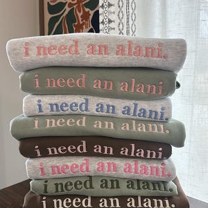 May include: A stack of eight green, gray, and brown sweatshirts with the phrase "I need an alani." embroidered in pink, white, and blue on each sweatshirt.