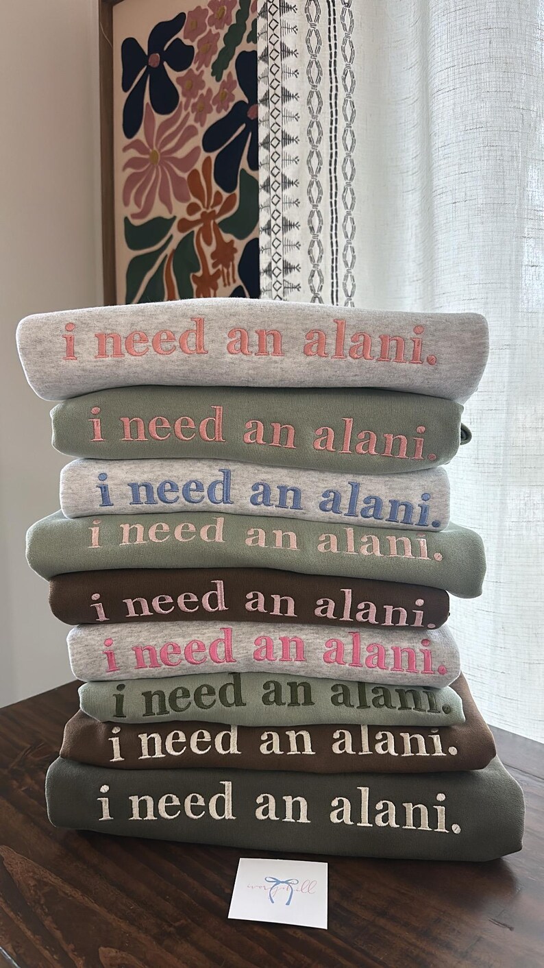 May include: A stack of eight green, gray, and brown sweatshirts with the phrase "I need an alani." embroidered in pink, white, and blue on each sweatshirt.