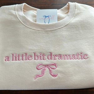 May include: A cream-colored sweatshirt with the text "a little bit dramatic" embroidered in pink. A pink bow is embroidered below the text.
