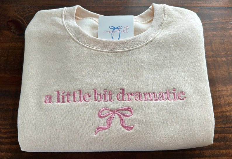 May include: A cream-colored sweatshirt with the text "a little bit dramatic" embroidered in pink. A pink bow is embroidered below the text.