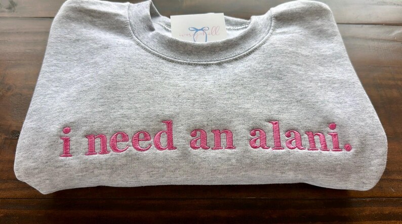 May include: A gray sweatshirt with the phrase "i need an alani." embroidered in pink.