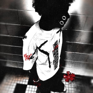 May include: A white t-shirt with black and red graphics. The shirt has a design on the sleeve and the front. The image is in black and white with red accents. The person is wearing black pants and white shoes.