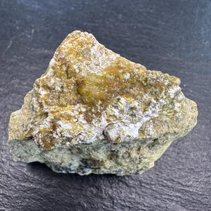 May include: A raw, triangular-shaped mineral specimen featuring a textured, earthy green and tan exterior. The surface displays a mix of matte, sandy-colored rock and patches of translucent, amber-colored crystalline formations with a reflective luster.