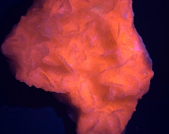 May include: A vibrant orange mineral specimen with a layered, textured appearance. The mineral has a glowing, almost fluorescent quality, with small, sparkling inclusions. The background is a deep, dark blue, creating a striking contrast.