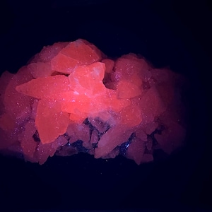 May include: A cluster of bright, glowing orange crystals against a dark background. The crystals have a rough, irregular texture and appear to be illuminated from within, creating a striking visual effect. The overall composition suggests a mineral specimen.