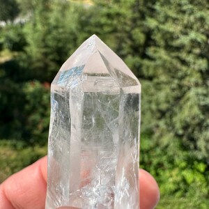 May include: A clear quartz crystal point, held in a hand, with a natural, faceted shape. The crystal is transparent, allowing light to pass through, and has a polished, smooth surface. The background is blurred, showing green foliage.