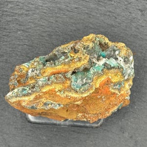 May include: A rough-hewn mineral specimen with a mix of colors. The rock features areas of teal, orange, and brown, with a crystalline texture. The mineral sits on a clear plastic stand, showcasing its natural beauty. This unique piece is ideal for collectors.