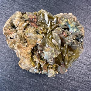 May include: A natural mineral specimen with a rough, irregular shape. The stone features a mix of colors, including shades of green, brown, gold, and iridescent hues. The surface has a crystalline structure, with visible formations and textures.