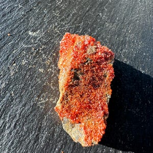 May include: A striking orange mineral specimen with a crystalline structure. The mineral has a rough, textured surface and is set against a dark, textured background. The colour is a bright, saturated orange, with hints of darker shades in the crevices.