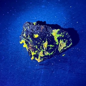 Puede incluir: Una roca oscura de forma irregular con vibrantes parches fluorescentes amarillos y verdes. La roca se encuentra sobre un fondo azul oscuro, creando un contraste llamativo. La textura parece áspera y desigual.