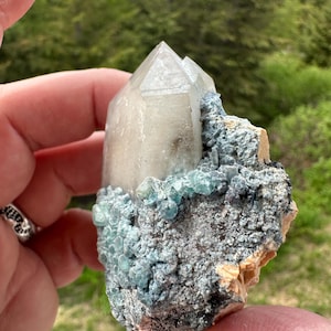 May include: A raw mineral specimen featuring a large, translucent, pointed quartz crystal cluster emerging from a textured matrix. The base is covered in small, vibrant teal-coloured crystalline formations and rough, greyish-brown rocky material.